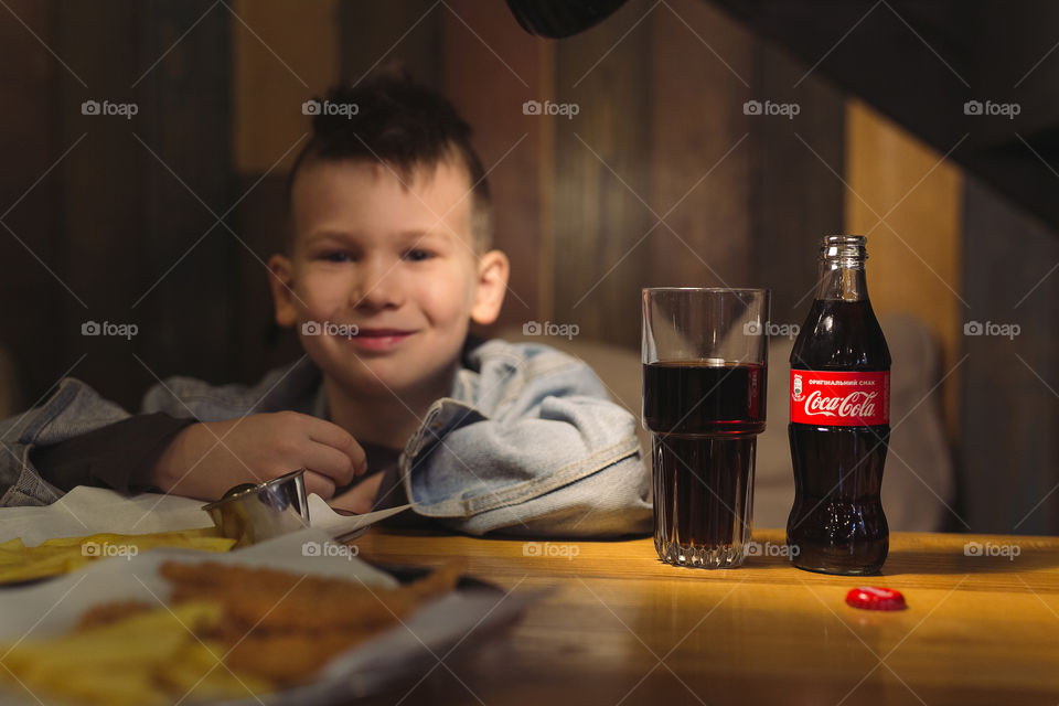 cute kid is sitting in a restaurant, having lunch and drinking Coca-Cola.  boy wearing denim clothes