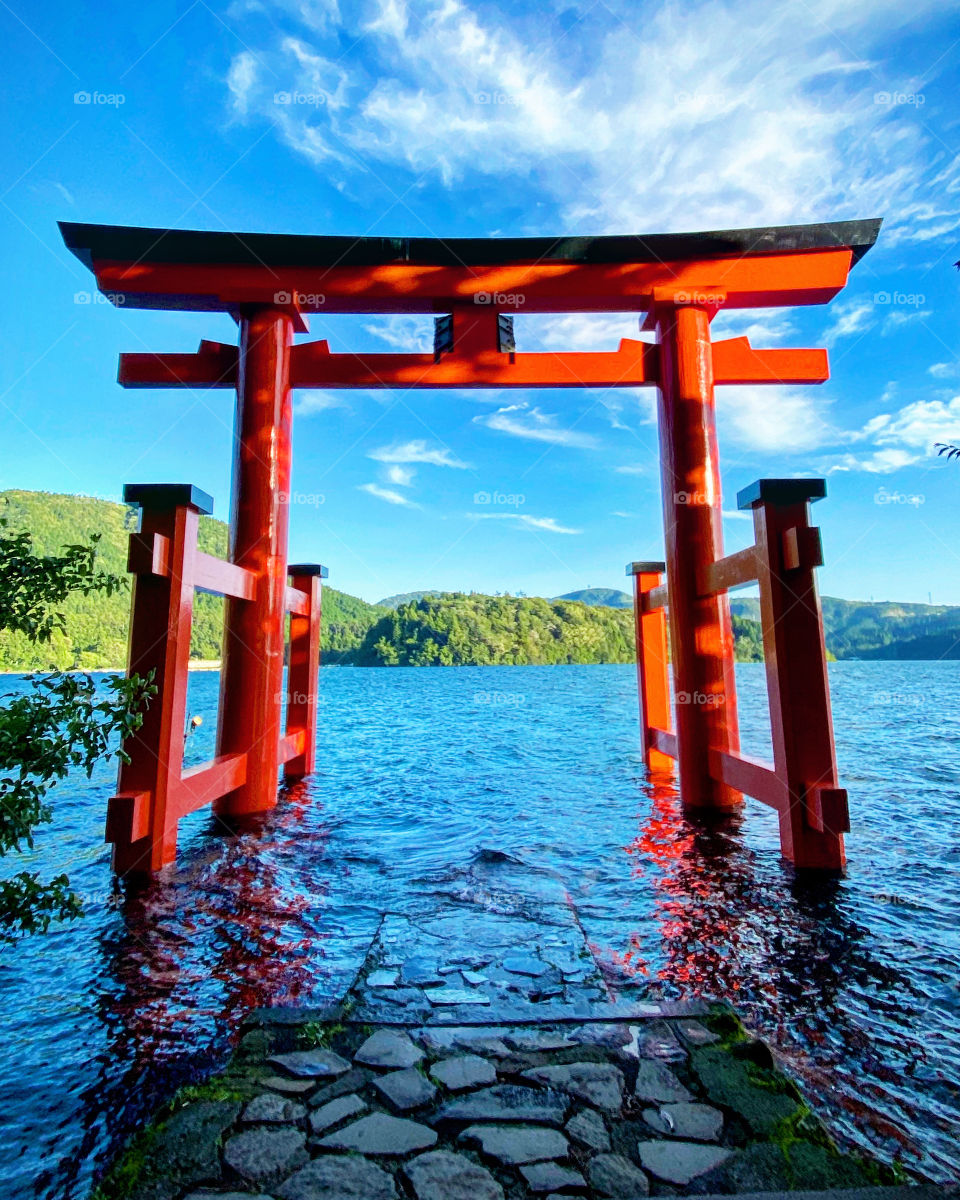 Large red torii gate set inside a lake, water swirling against a blue sky, dragon-like clouds and greenery on the other side of the lake. Hakone, Japan