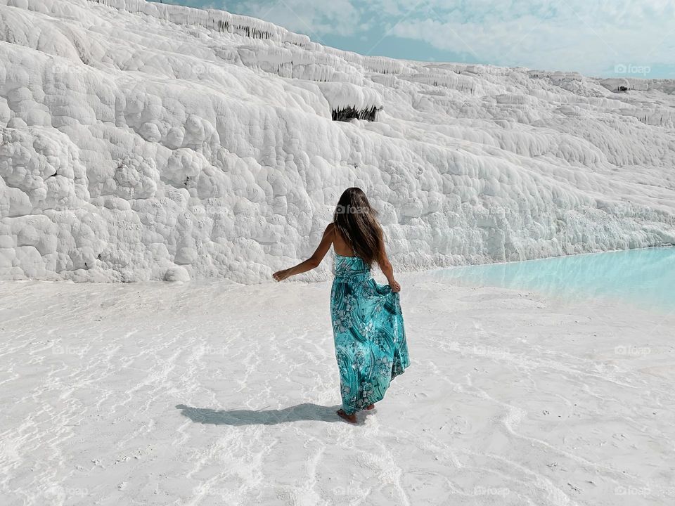 Young woman with long hair in blue dress walking by white chalk mountain with blue water 