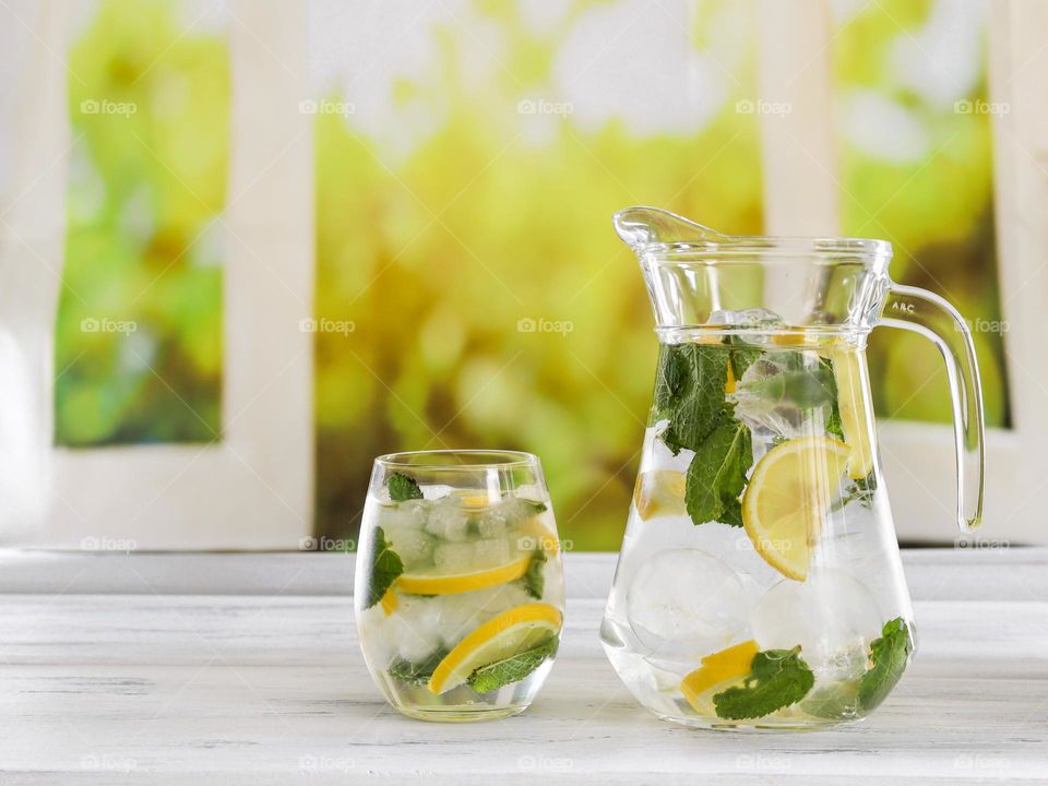 Cold refreshing lemon drink with mint large ice balls in a decanter and a glass standing on a light wooden shabby table against the background of a blurred window with a green flowering field, side view cope plan. Summer drinks concept.