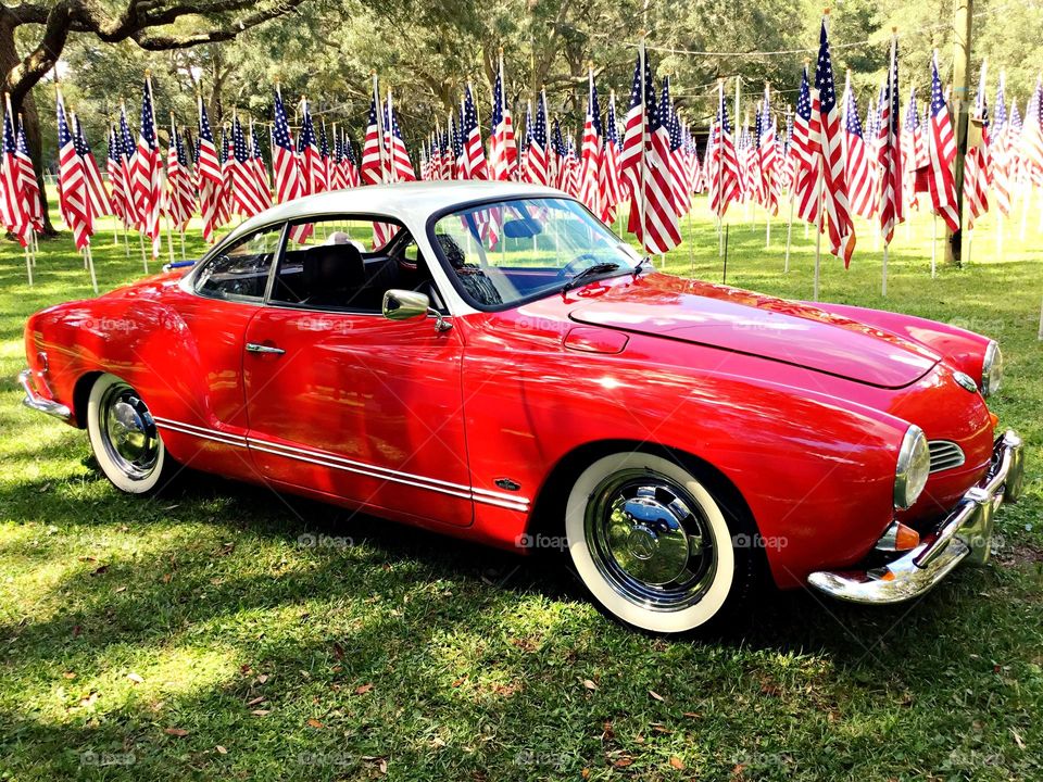 A shiny Red and white Karmann Ghia, Volkswagen with chrome hubcaps rests in the shade surrounded by several US Flags