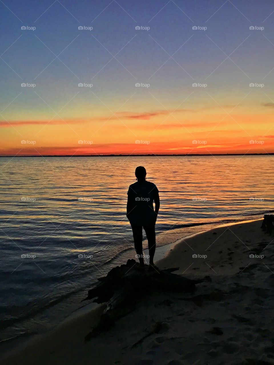 A young lady strolls down the waters edge during sunset. 
The big orange ball slowly goes down and down and it sets into the horizon making the sky absolutely stunning.