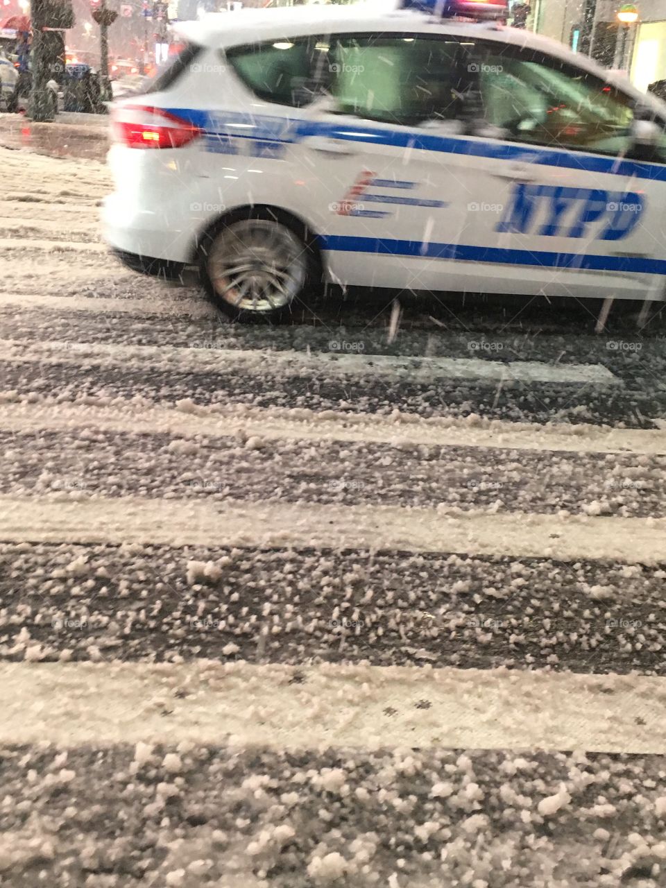 A picture of the pavement on a snowy evening with a New York City police car driving past 