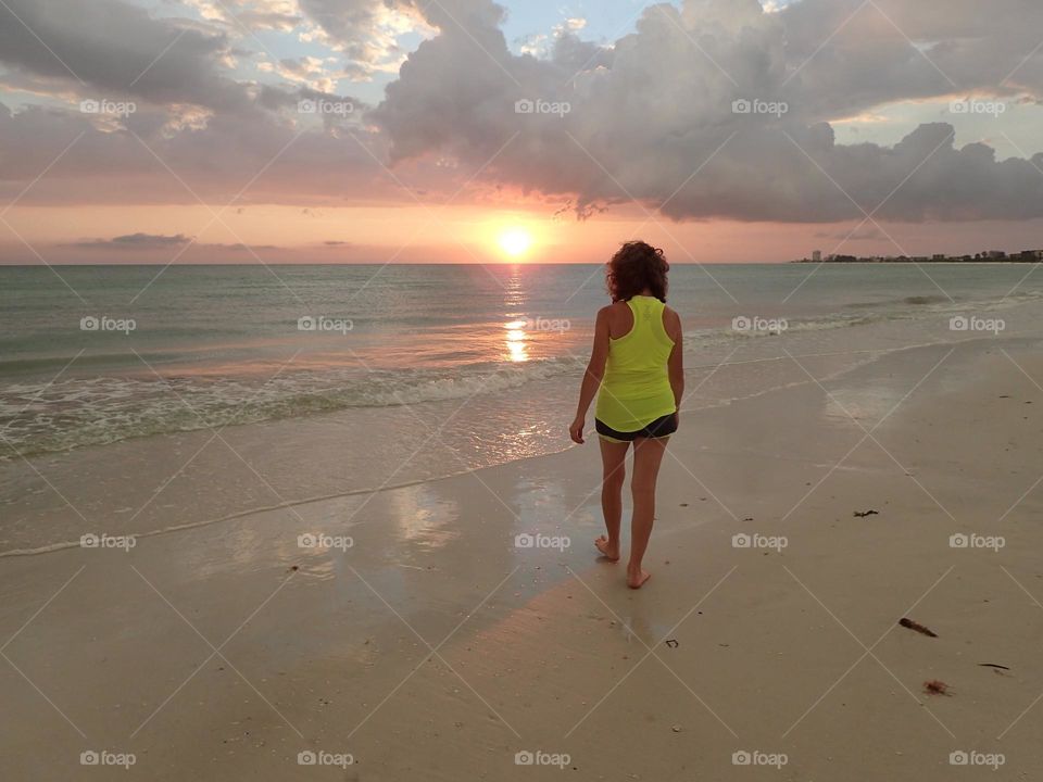Unusual pastel colored beach scene of Woman walking towards sunset full of Dark clouds with silver lining on horizon