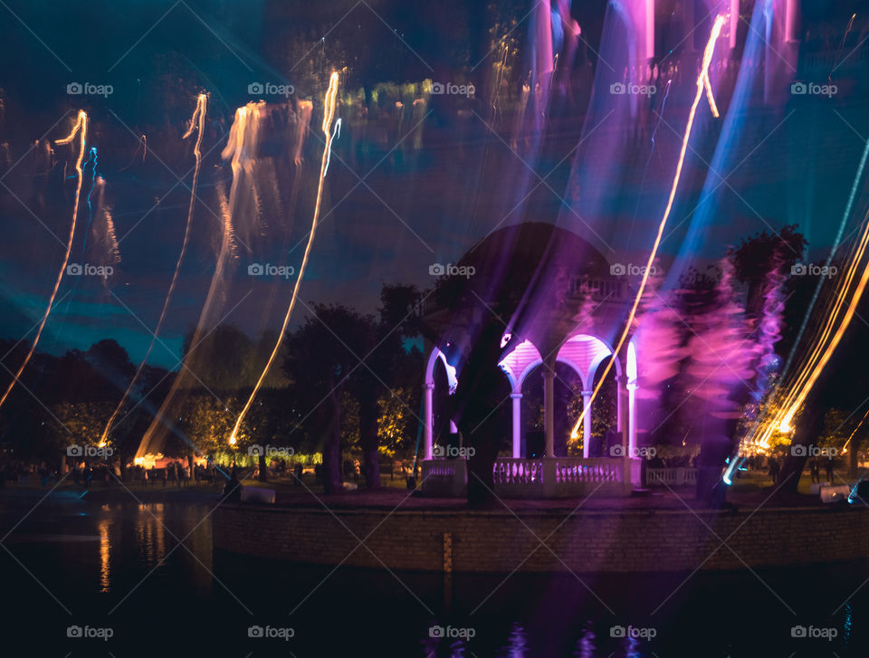 Night view of a lit up bandstand in a pond with colourful lighttrails.