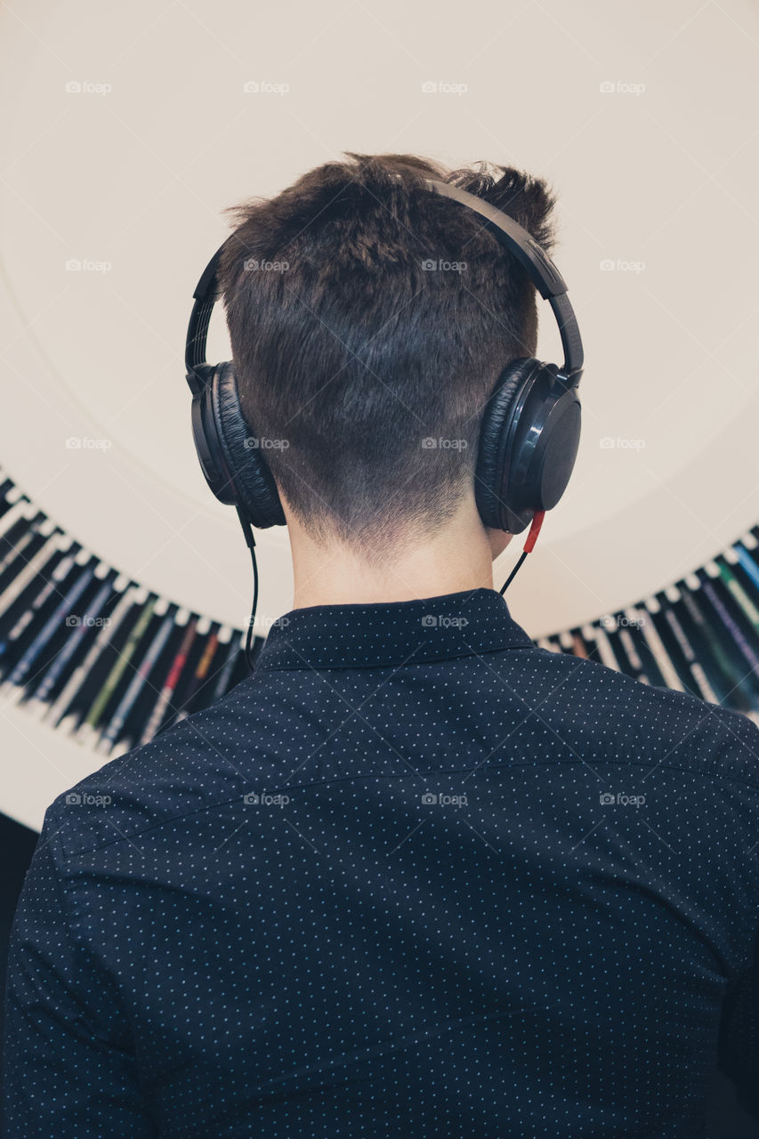 Young man listening to music through headphones standing next to bookshelf with recordings