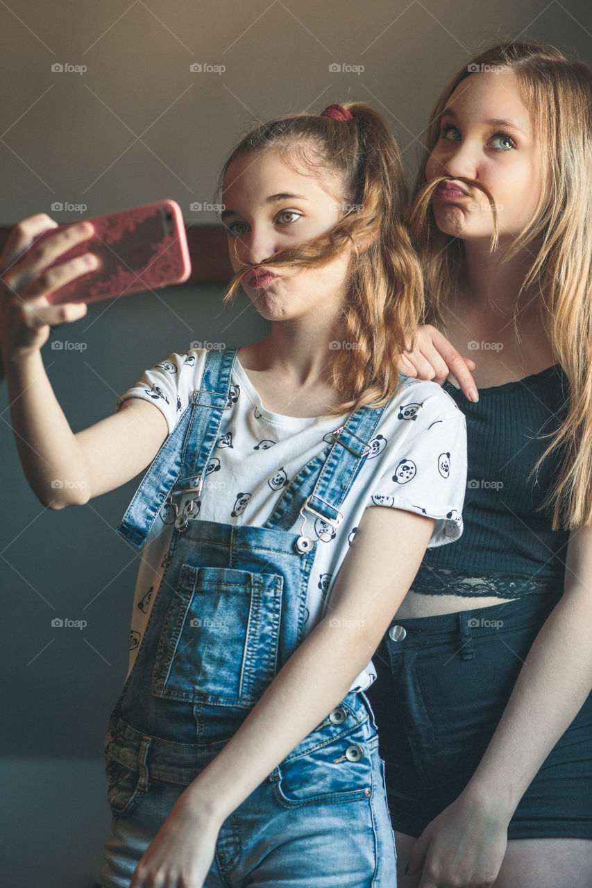 Young women taking selfie, using smartphone camera. Girls making faces, enjoying taking funny pictures together