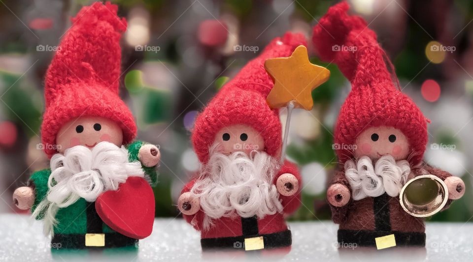 3 tiny, white bearded Christmas Folk made of wood and fabrics, standing on a glittery surface with a blurred background of lights and decorations 