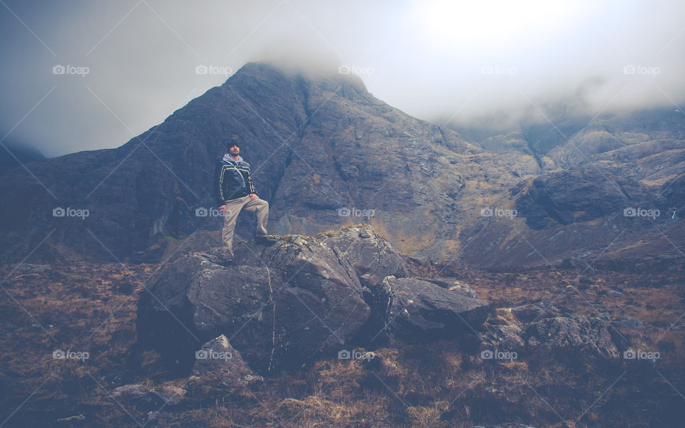 A man stands upon a large bolder, as mist swirls around the area, high in the mountains on the Scottish Isle of Skye.