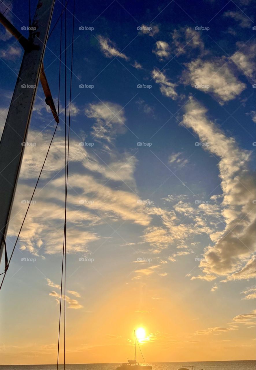 Basking in the Caribbean sunlight, a solitary boat floats idly on the azure sea. The sun, a radiant orb, mirrors a pathway of shimmering gold on the water's surface, creating an image of serene tranquility.