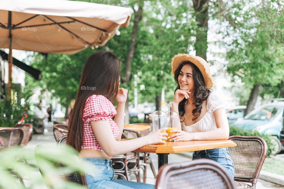 Happy smiling beautiful brunette young women friends in summer clothes with juice in hands sitting in cafe at the summer city street