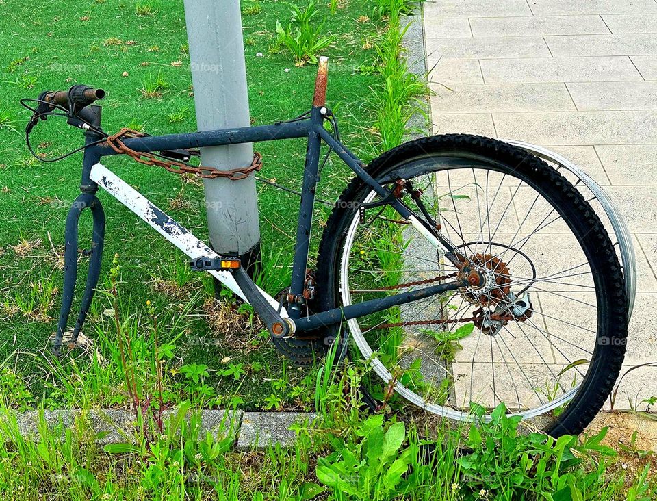 Transformer and terminator bicycle. On the green grass there is a bicycle tied with a metal chain with a lock to a post. The bike does not have a steering wheel, seat and rear wheel