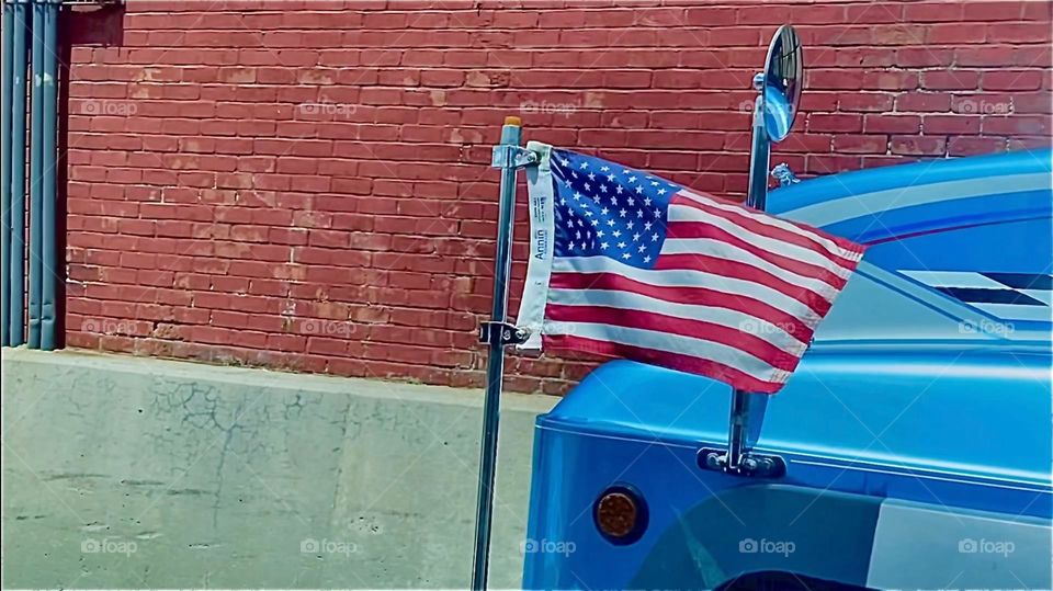 Another image in a series of stills showing the movement of the red white and blue „American flag“ attached to an unusual looking blue metallic car as it is blown about by the brisk wind causing it to undulate wildly. 2022. Hypnotic Productions