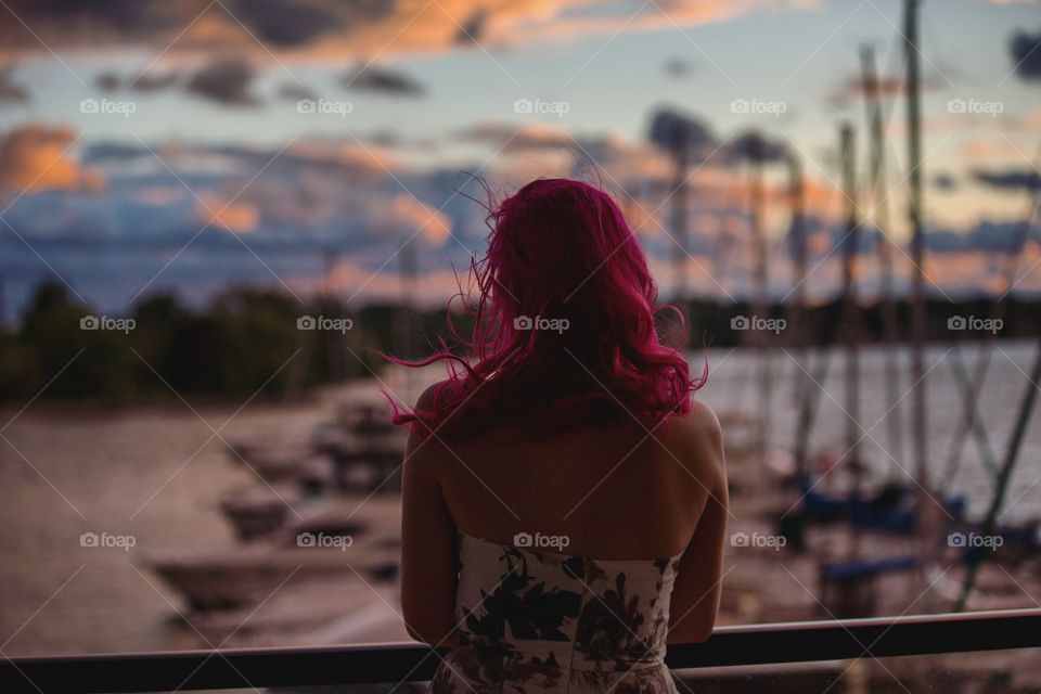 A young woman with pink hair overlooking a harbour at sunset