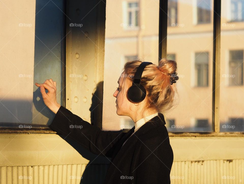 Young blonde woman in headphones standing near window in sunny day, portrait of woman.