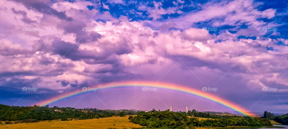 Landscape with beautiful Rainbow and clouds with pink and purple tones, contrasting with the blue sky.
Paisagem com lindo Arco-íris e nuvens com tonalidades rosa e roxo, contrastando com o céu azul.