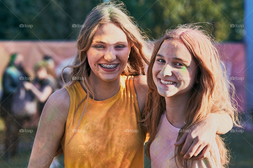 Portrait of happy smiling young girls with colorful paints on faces and clothes. Two friends spending time on holi color festival. Real people, authentic situations