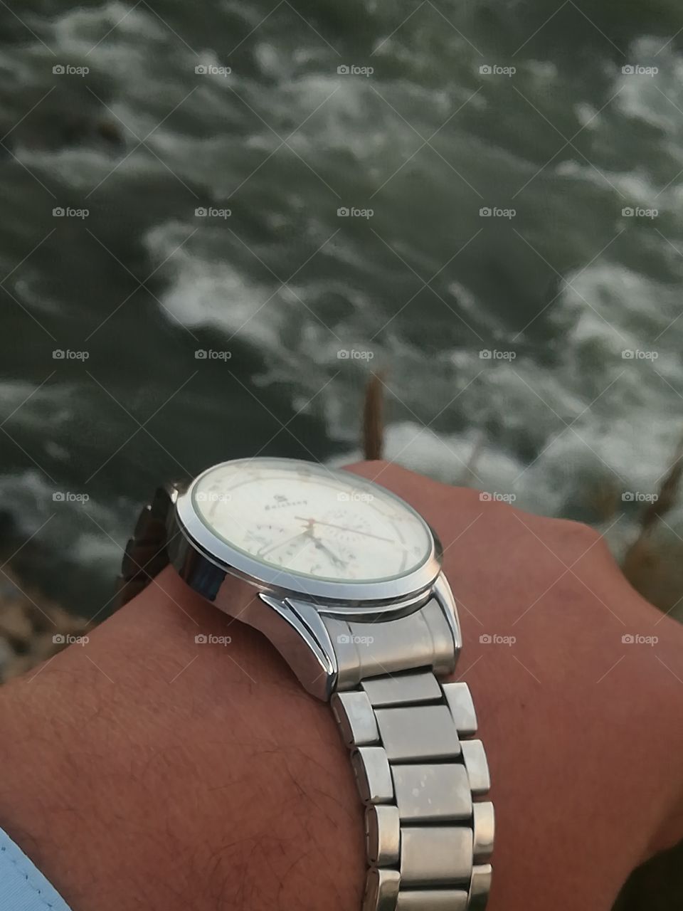 The wrists watch on mans wrists in front of the river bank.