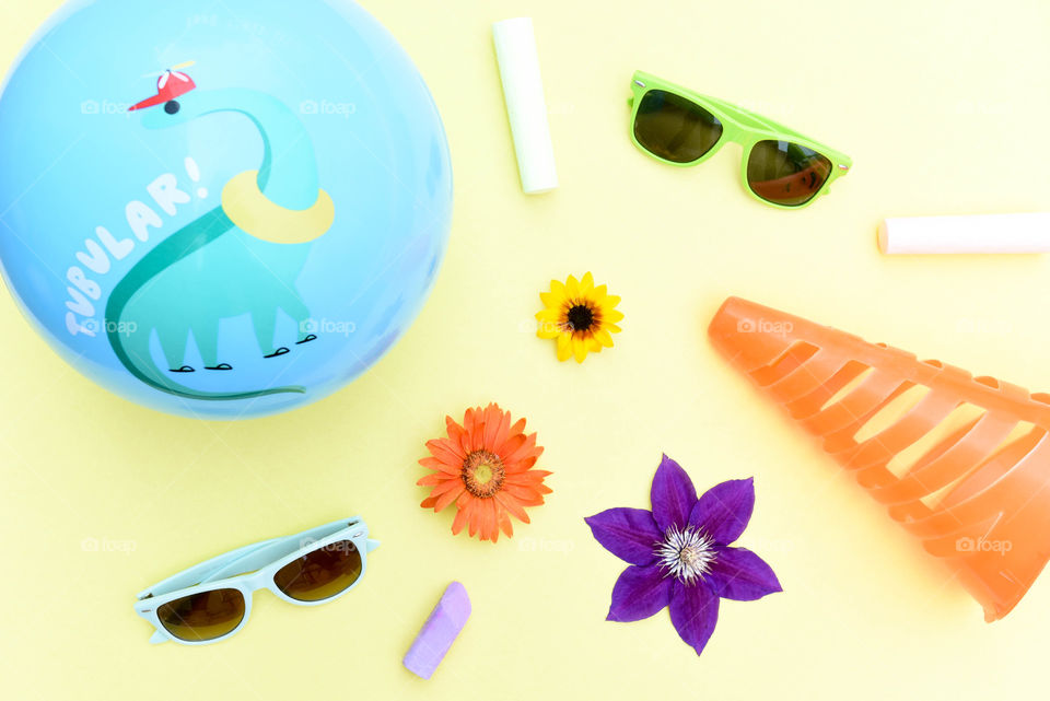 Flat lay of colorful summer objects including sidewalk chalk, sunglasses, flowers and a ball