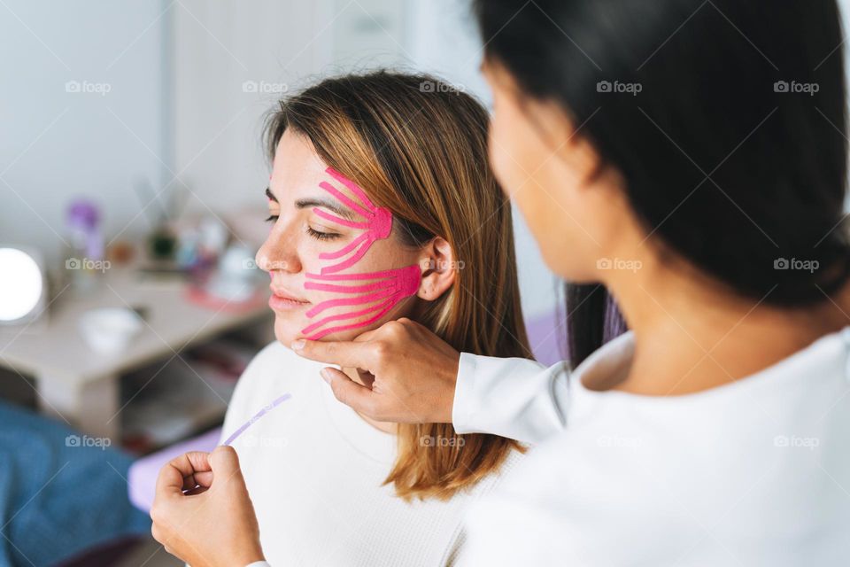 Young beautiful brunette woman with long hair doctor cosmetologist makes face taping for patient in office. Young women do anti-aging procedures with help of kinesio tape