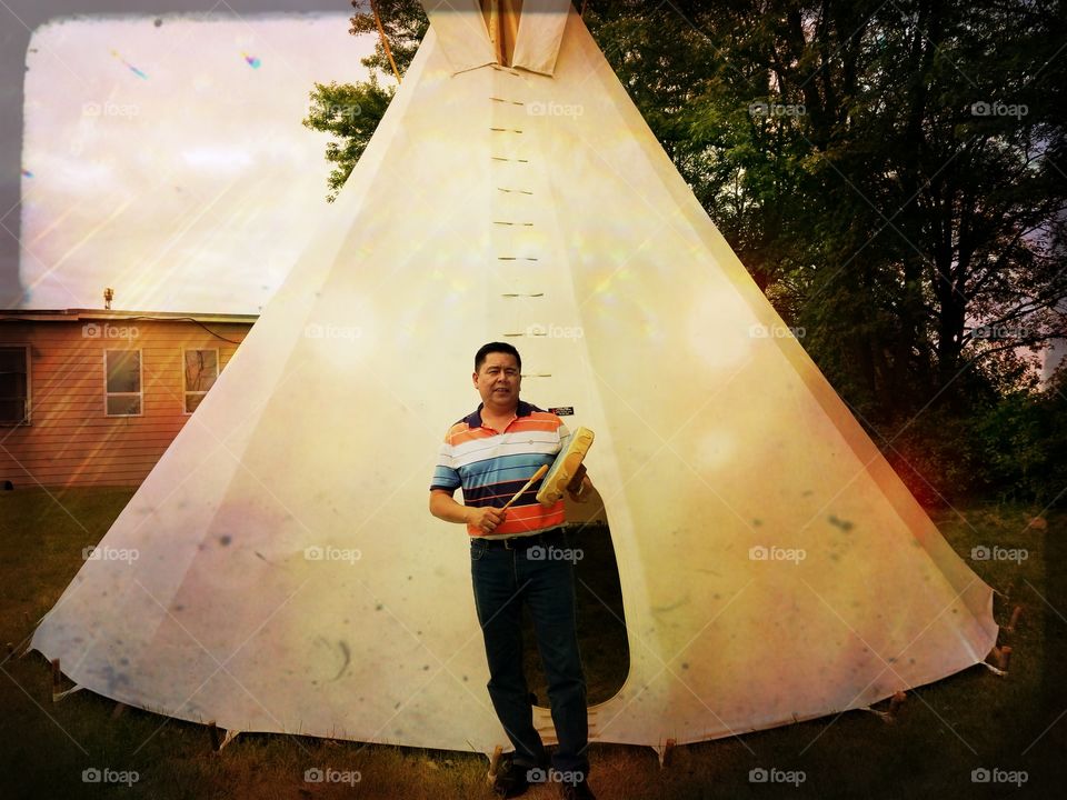 First Nations drummer stands in front of a teepee.