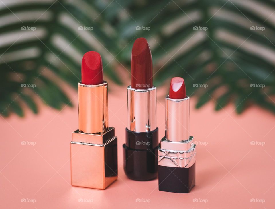 Three red lipsticks stand on a pink background with blurred back branches of a palm tree on a white brick wall, close-up side view. The concept of female cosmetics, beauty salon.