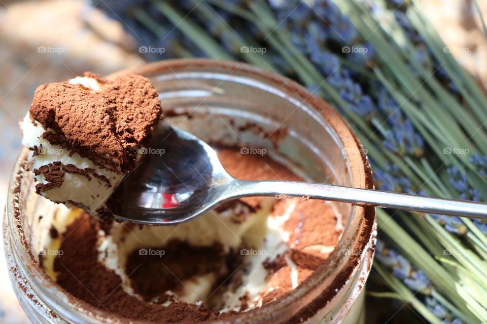 Tiramisu in a glass jar, small heart shaped piece in a spoon - ready to be eaten... on the background lavender laying on a table 