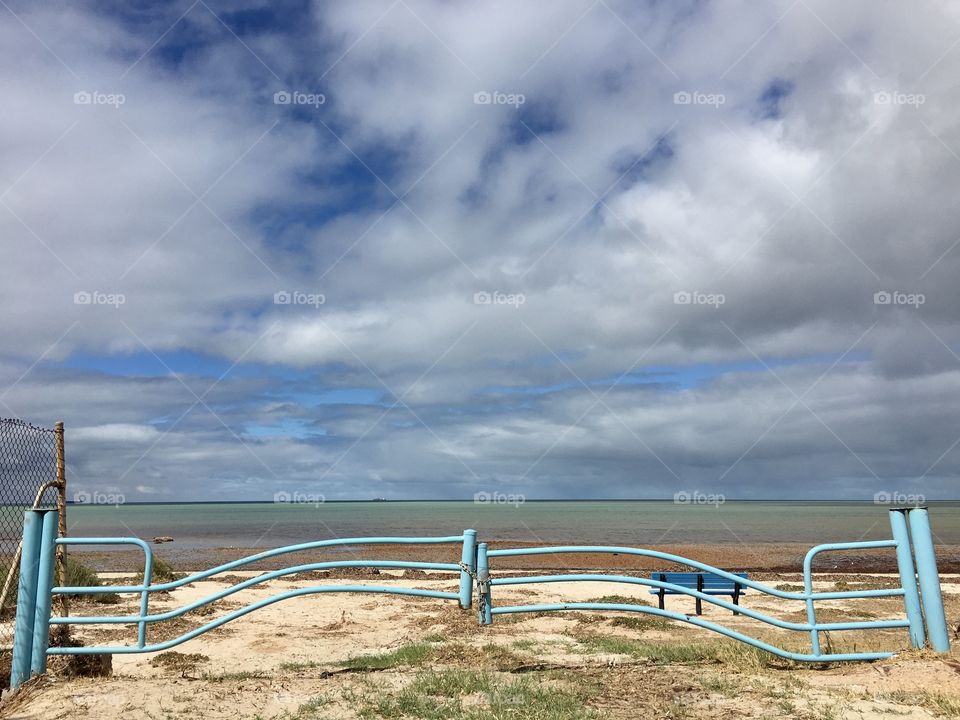 Dreamy aqua marine blue metal gate padlocked and locked at approach to ocean beach with blue bench, ocean, beyond it and billowy cloudy sky