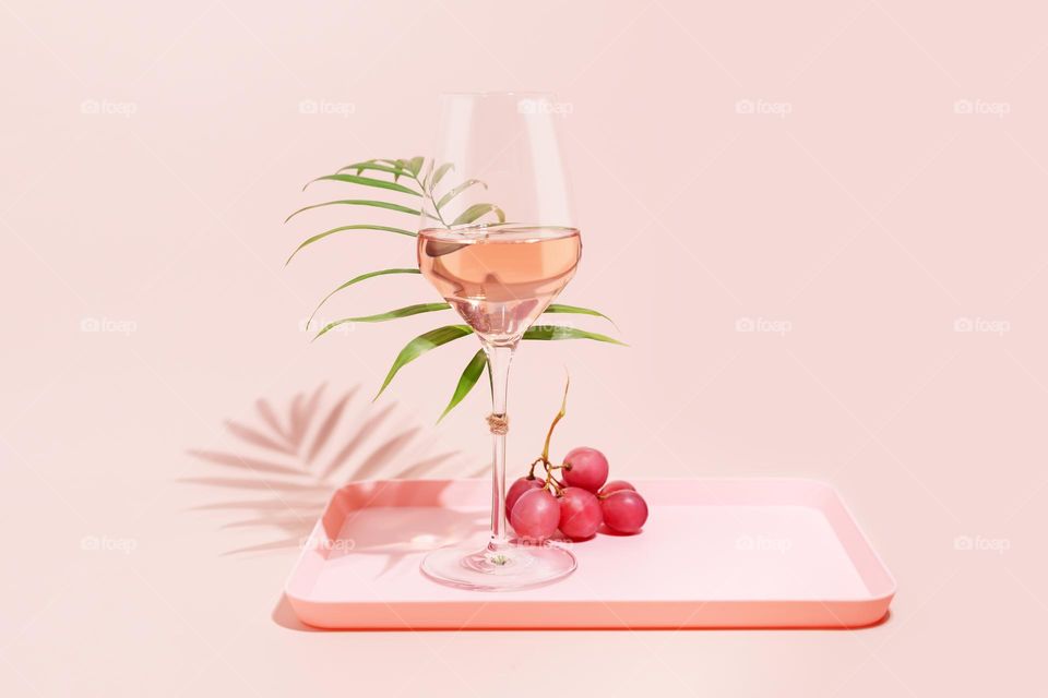 Rose wine. Minimalistic, Summer, holiday composition with glass of rose wine, grapes and palm leaf on a pink tray on a pink background