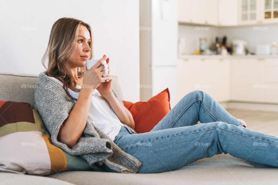 Young beautiful woman forty year with blonde long curly hair in cozy knitted grey sweater with cup of tea in hands in bright interior at the home