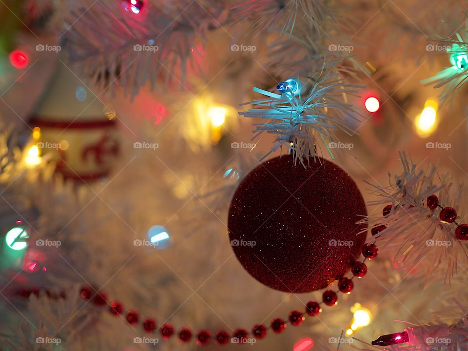 A red glitter ball ornament hanging from a branch of a white Christmas tree amongst the soft glow of lights. 