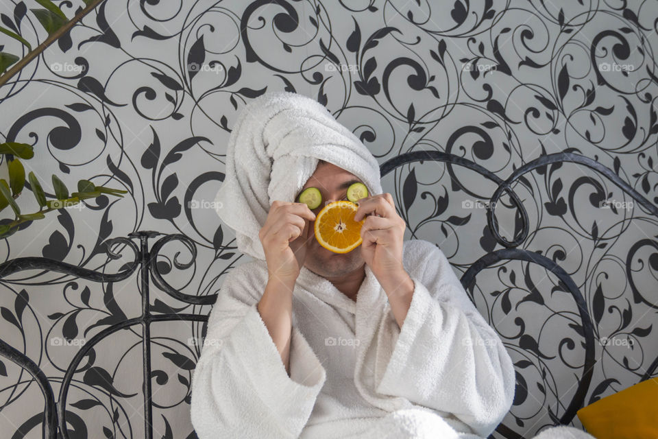 A man and home spa care at home takes care of himself and smears his face with cream, does a manicure at home on a blue background and with oranges and rests.