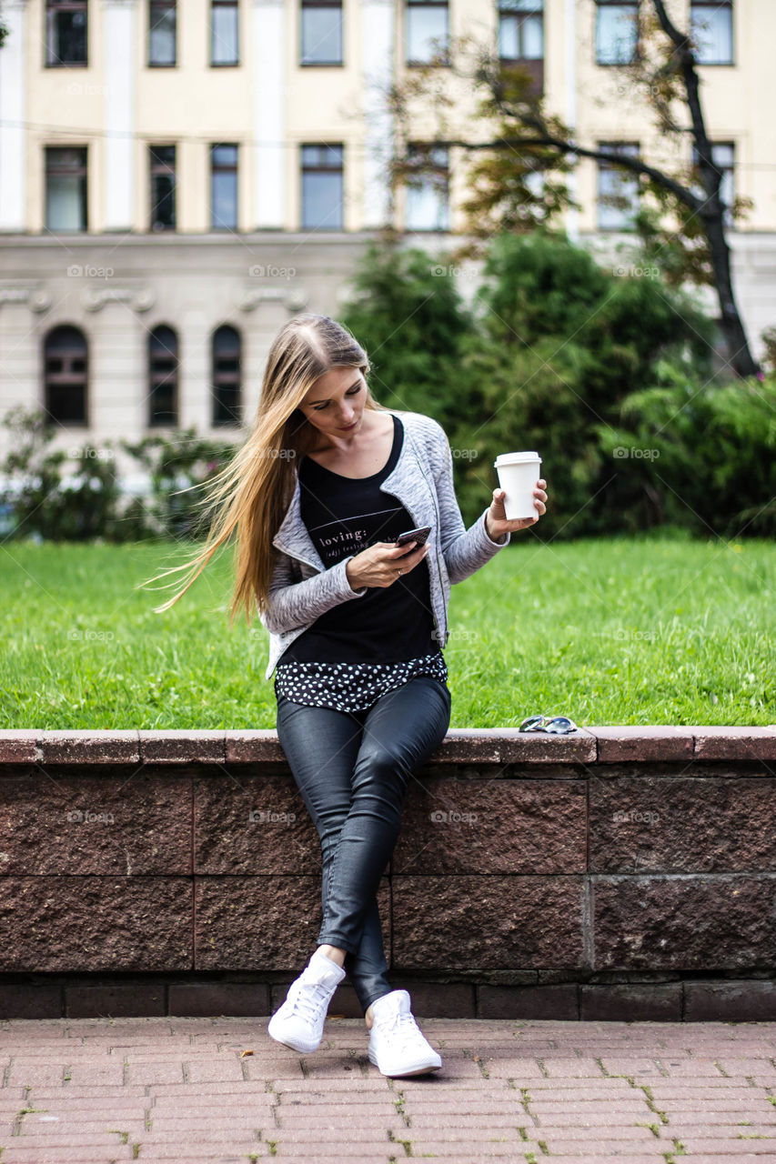 Woman using mobile phone holding drink against building