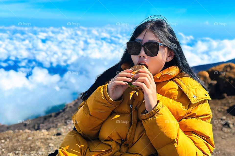 Portrait of young woman enjoying burger on mountain top above beautiful clouds