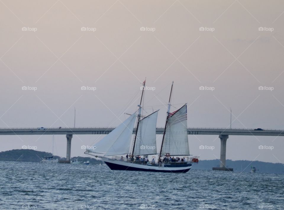 A large sailboat with 3 sails on a choppy river in front of a bridge at dusk