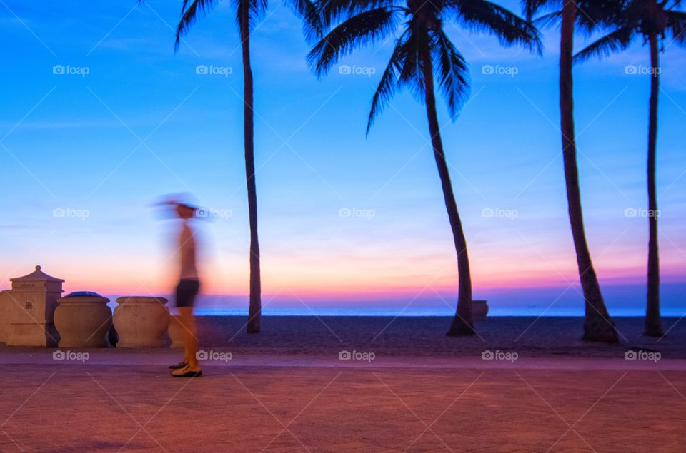 A walker is blurred as the sun creates silhouettes of the palm trees on an early morning sunrise at Hollywood Beach, Florida.  