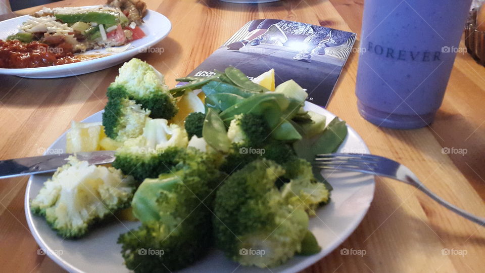 A nutrition photo. The focus on the broccoli and the plate in general tells us "This is what I'll eat today". The sports booklet in the background says "I'm planning to also do sports today". Also, in the background there is another plate with a delicious meal on it which can be perceived as a contrast to what we see in the forefront.