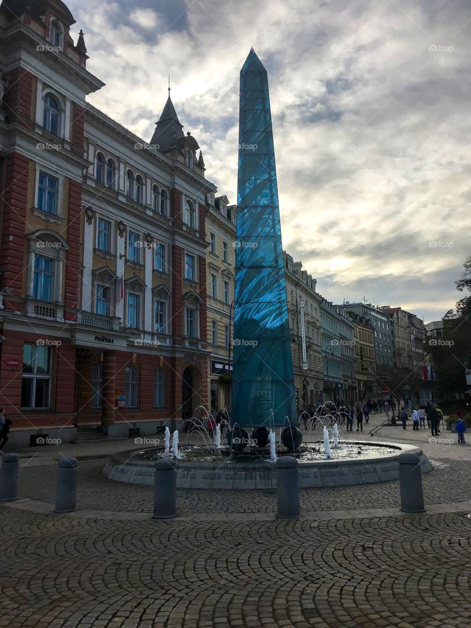 A pillar in the middle of the fountain , a series of buildings of different colors .There are many people walking in the distance, the sky was cloudy, with the golden glow