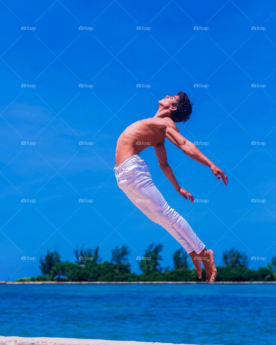 Portrait of a person jumping near the beach with a bright blue sky and calm sea water in the background