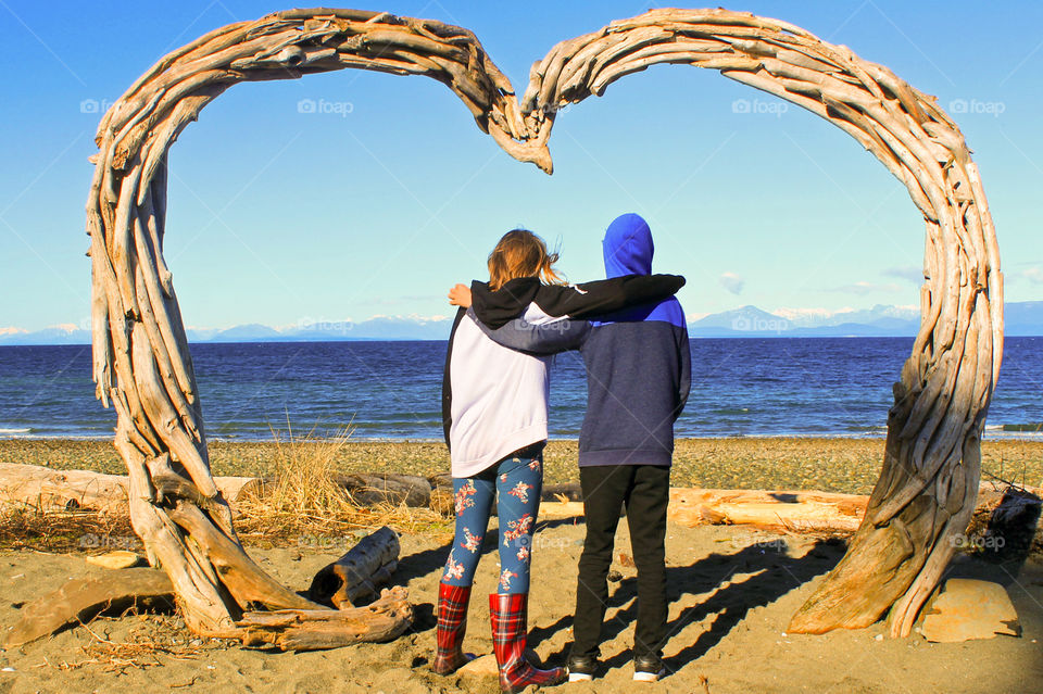 Symmetry of a driftwood heart with the photo centred where the two curves meet above the two friends. They share a moment gazing at the beautiful Coastal mountains across the Salish Sea as the golden sun begins to set behind them. 