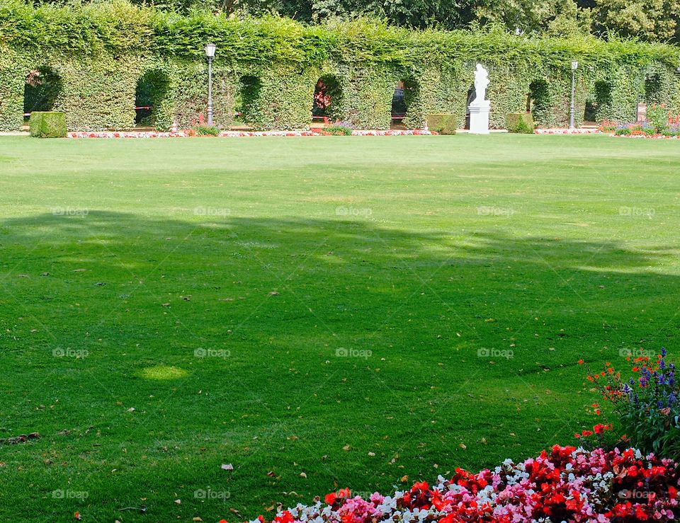 A large open grassy area with bright flowerbeds in the foreground and statues, hedges, and lampposts in the background on a sunny summer day in an urban park in Europe. 