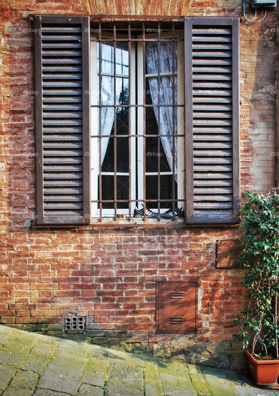 Lace curtains and soccer shoes. Soccer shoes set perched atop windowsill backed by lace curtains call and brown shutters in the village of Sienna