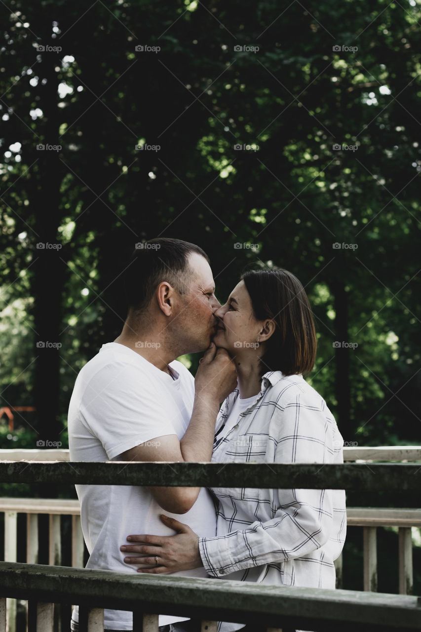 One young beautiful couple are standing on a wooden old bridge in a public park and a man is kissing a happy woman holding her chin with his hand, close-up side view. The concept of kissing people.