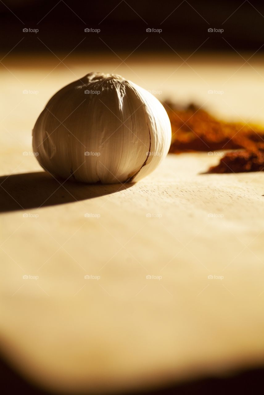 A single garlic clove rest on a wooden cutting board with a variety of herb spices.