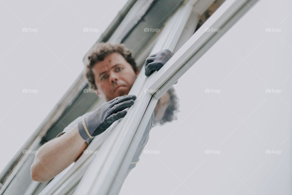 A young Caucasian man wearing construction gloves looks out of a window repairing a window frame, close-up view from below with selective focus. Concept people and windows .