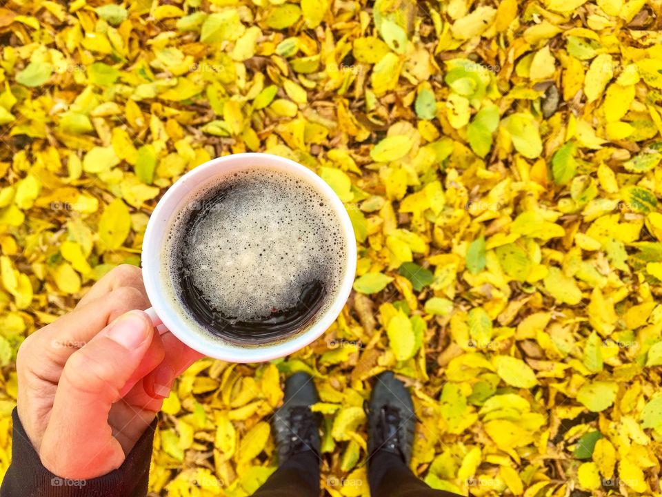 Hand holding cup of coffee looking down at yellow leaves fallen on the ground