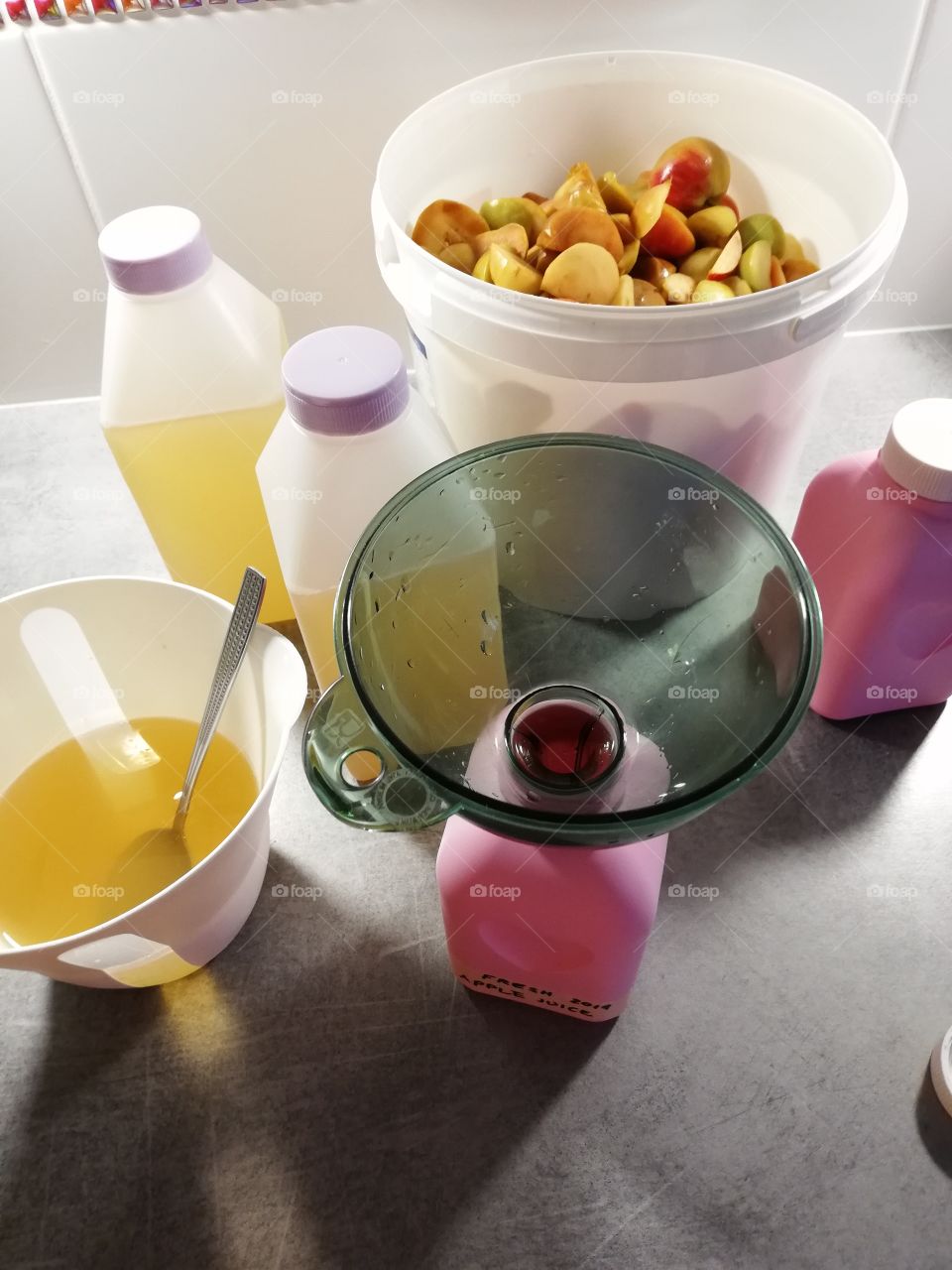 Homemade apple juice is done and is in plastic bottles. Some yellow juice and a tablespoon are in a jug. In a white bucket halves of fruits. A green funnel in a pink bottle, stoppers on the full bottles.