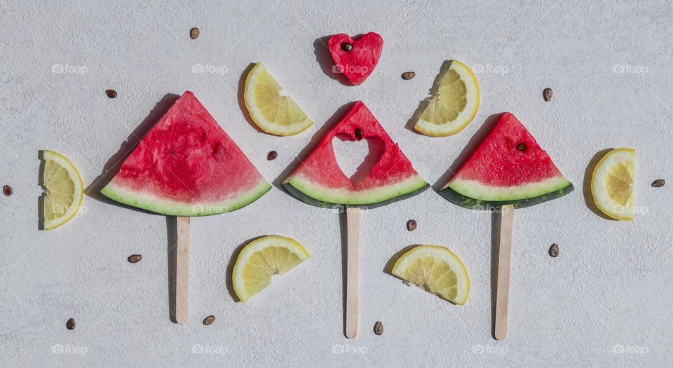 Three watermelon triangular pieces on wooden sticks with sliced ​​lemon and pits lie on a light background, flat lay close-up. Summer treats concept.