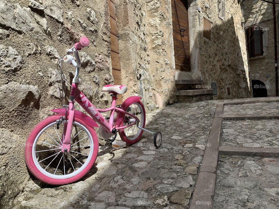 A pink children's bicycle leaning against the wall of an old house contrasts with the ancient world represented by the ancient town.