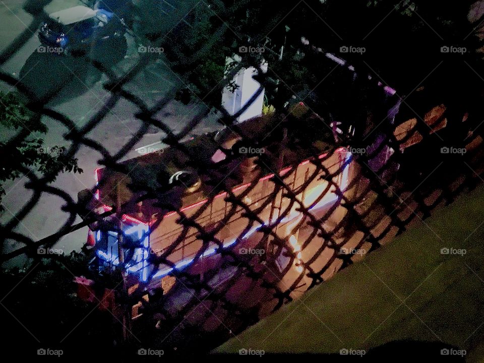 The illuminated takeout food vendor stand in the parking lot underneath the Pulaski Bridge is open 24 hours. This photo was taken in 2020 from the bridge looking down at the stand through the fence. Hypnotic Productions